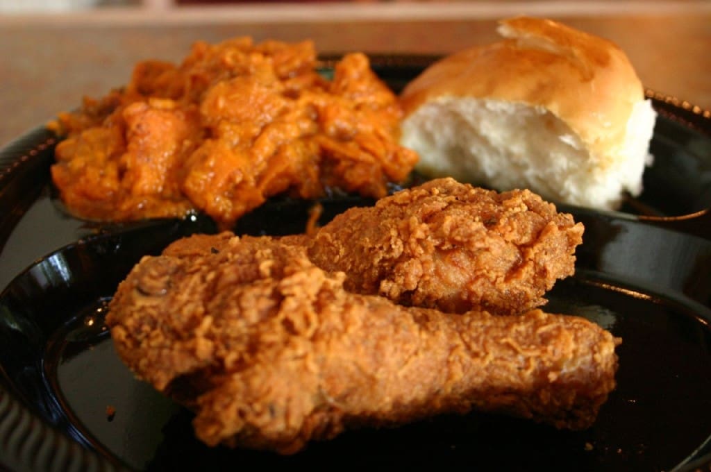 Close-up of a plate featuring two pieces of crispy golden-brown fried chicken, a serving of orange mashed sweet potatoes, and a soft white dinner roll.