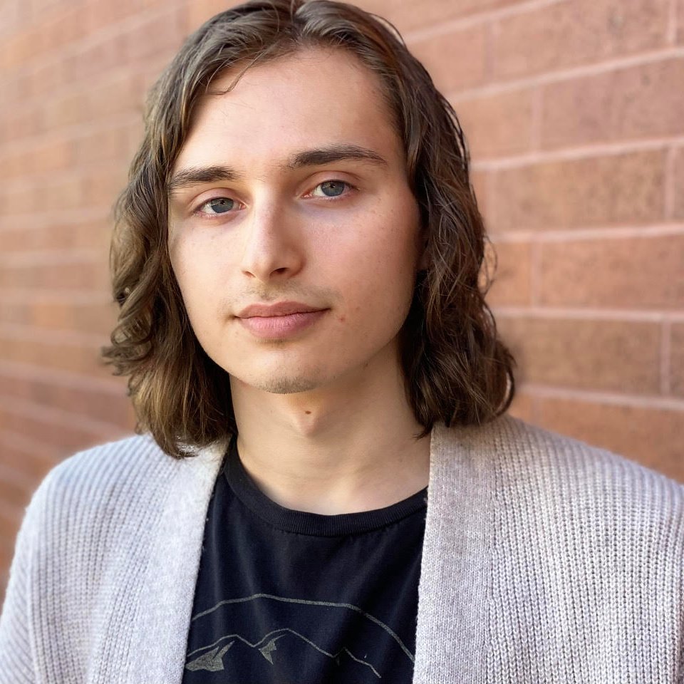 Aedan has shoulder-length brown hair and blue-gray eyes. They are wearing a light gray cardigan over a black tshirt and they are standing in front of a brick wall.