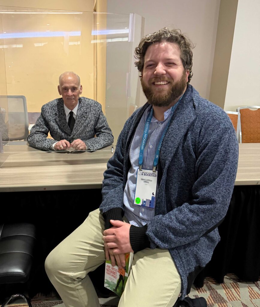 M.R. Lehman Wiens (foreground) sitting at a desk across from John Waters as they both smile at the camera. M.R. has short brown hair and beard and is wearing beige pants and a light blue button down shirt under a gray sweater. John is bald and wearing a gray suit and tie.