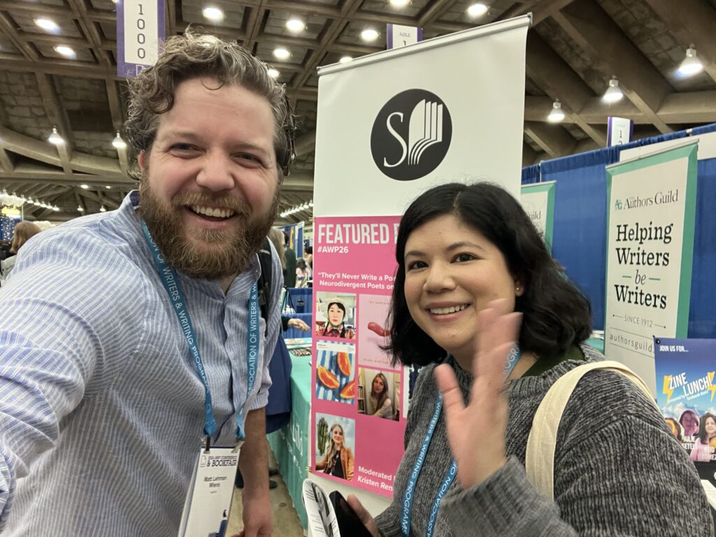 M.R. Lehman Wiens and Caitlin O’Halloran smiling at the camera. M.R. has short brown hair and beard and is wearing a light blue button down shirt. Caitlin has shoulder-length black hair and is wearing a gray sweater as she waves at the camera. They stand in the AWP book exhibit hall.