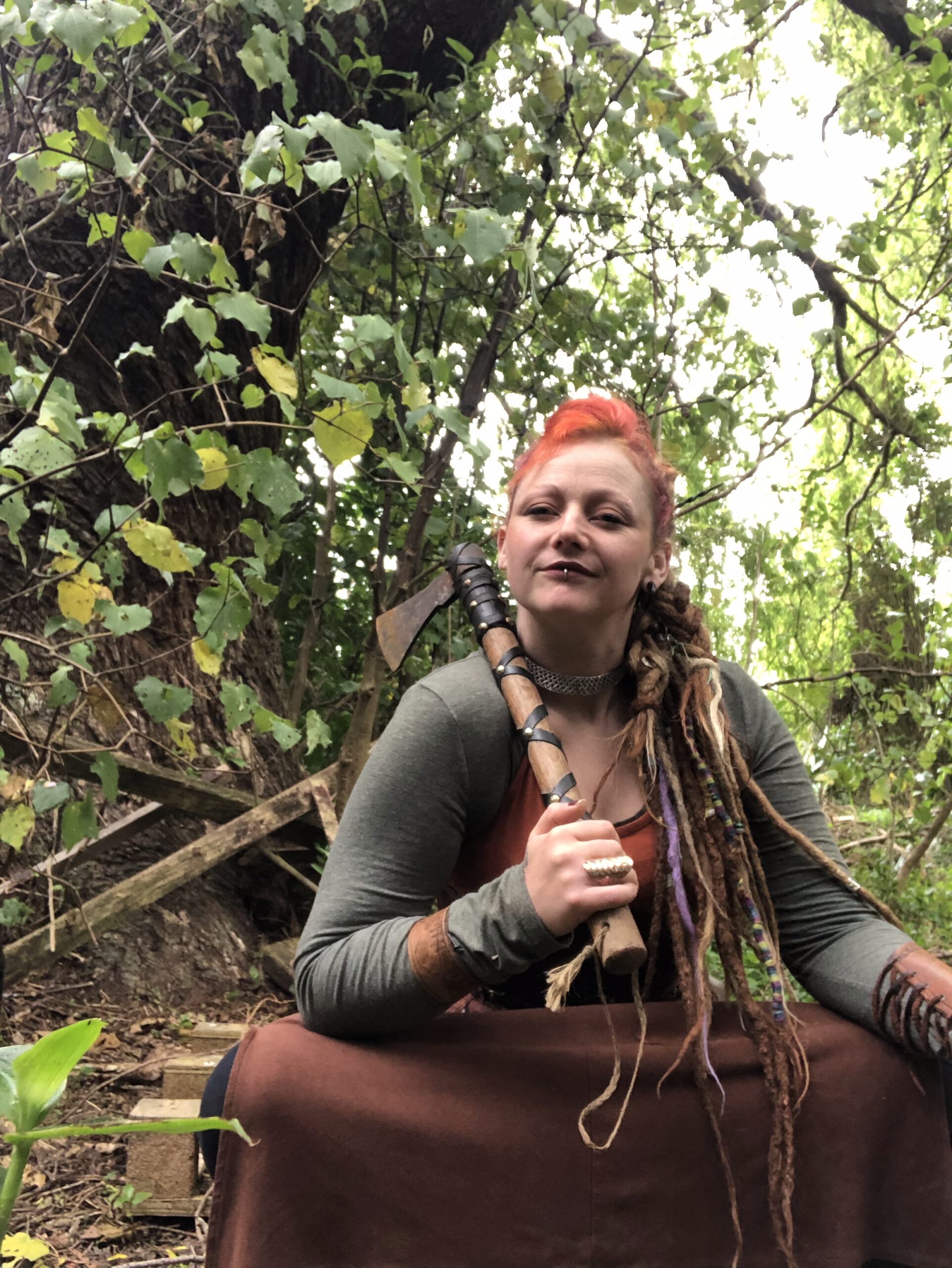 A striking outdoor portrait of Cerid in earth-toned attire, holding a hand-axe with a steadfast gaze. She is framed by the gnarled trunk of an ancient tree and lush, dappled greenery.