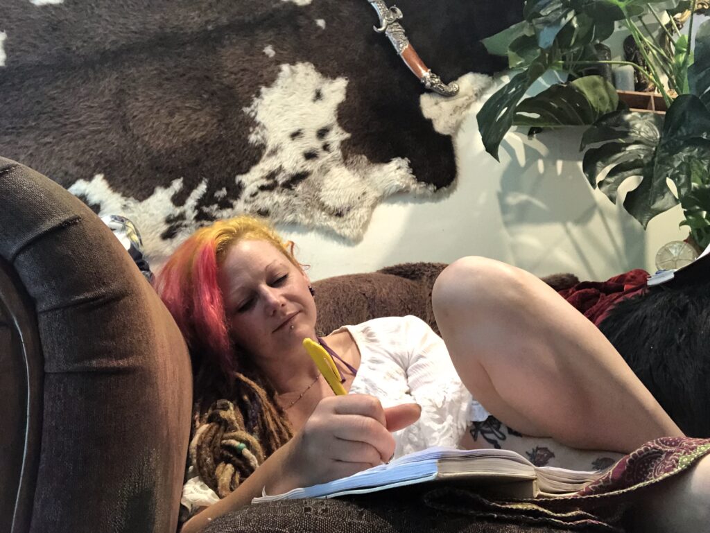 Cerid, with vibrant pink and yellow hair and long dreadlocks, is reclining comfortably on a brown couch while writing in a notebook with a yellow pen. Behind her, a cowhide rug hangs on the wall next to a decorative dagger and a lush Monstera plant, creating a warm and eclectic creative space.
