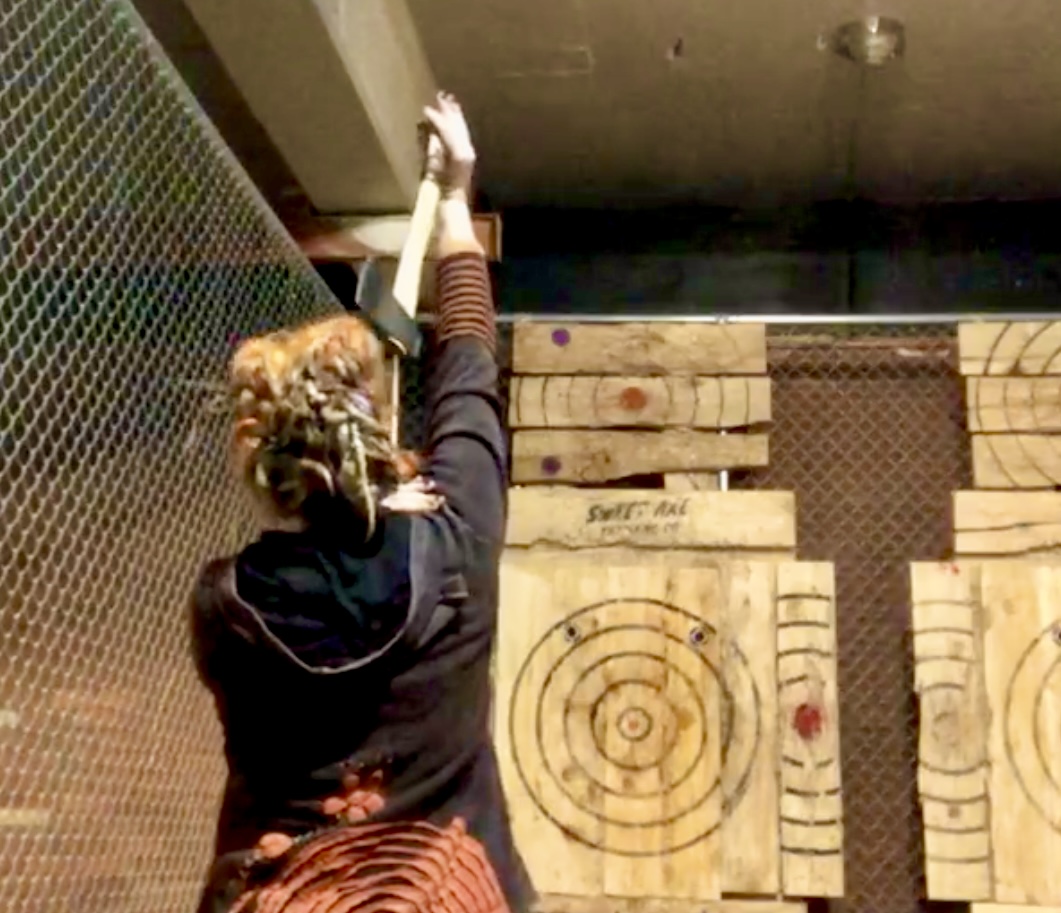 A photo from behind Cerid as she prepares to throw an axe at a wooden target. Her hair is styled in blonde and brown dreadlocks, and she is wearing a black hoodie with striped sleeves. She is holding the axe high above her head, aimed at a bullseye on a large wooden plank wall inside an indoor axe-throwing facility.