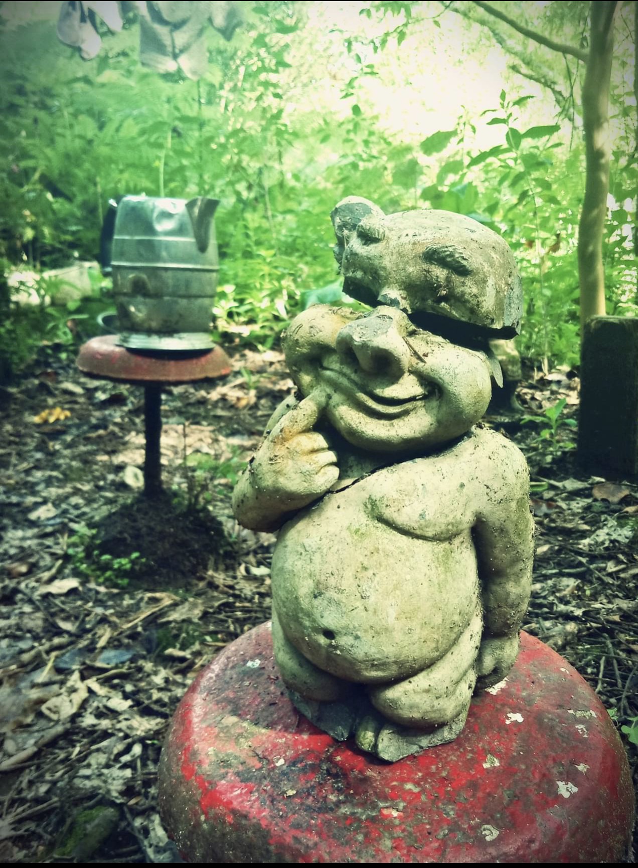 A weathered stone garden statue of a mischievous, smiling troll or gnome sitting on a red toadstool-style pedestal. The statue has a large crack across its head and is gesturing with a finger to its chin as if in thought. In the background, a lush green garden and a silver kettle atop another small stool are visible under soft, filtered sunlight.