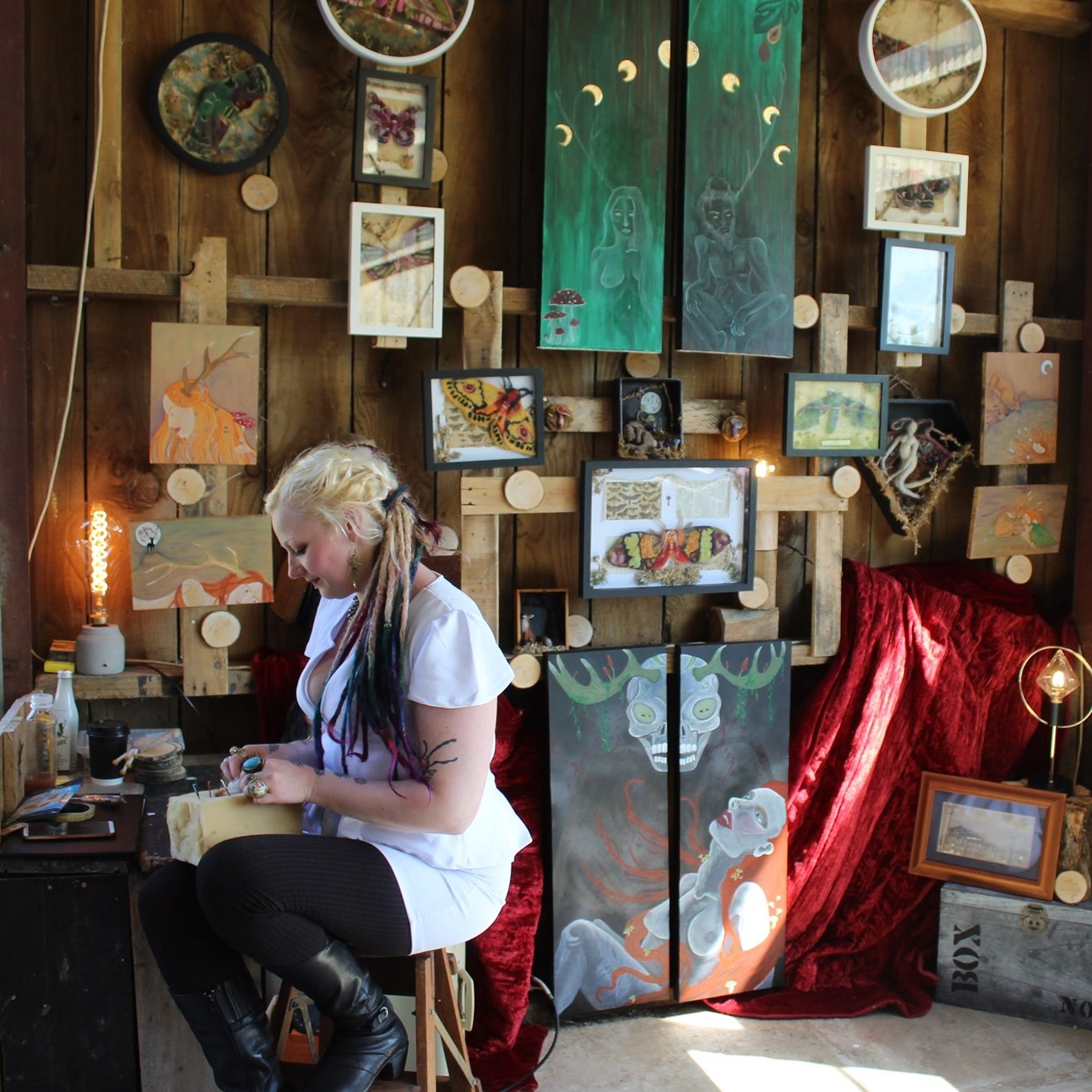 Cerid, with blonde braided hair and colorful dreadlocks, sits in her workshop carefully working on a craft project. She is surrounded by an eclectic gallery wall of her art, featuring paintings of ethereal figures with antlers, framed moth specimens, and moon phase illustrations. The space is warmly lit by an Edison bulb lamp, with rustic wooden walls and a red velvet drape adding to the cozy, artistic atmosphere.