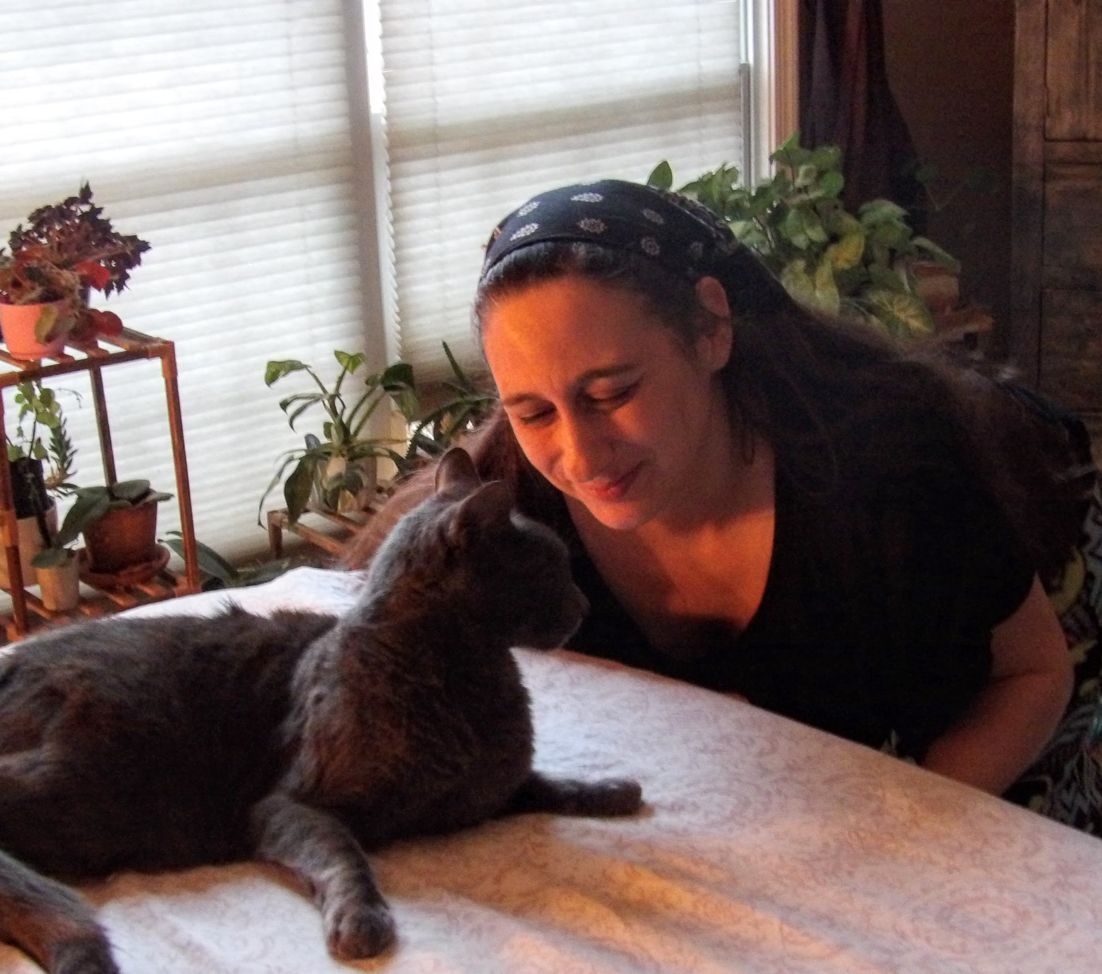 Mel, a woman with long dark hair, smiles down at her gray cat, Sam, with plants in the background.