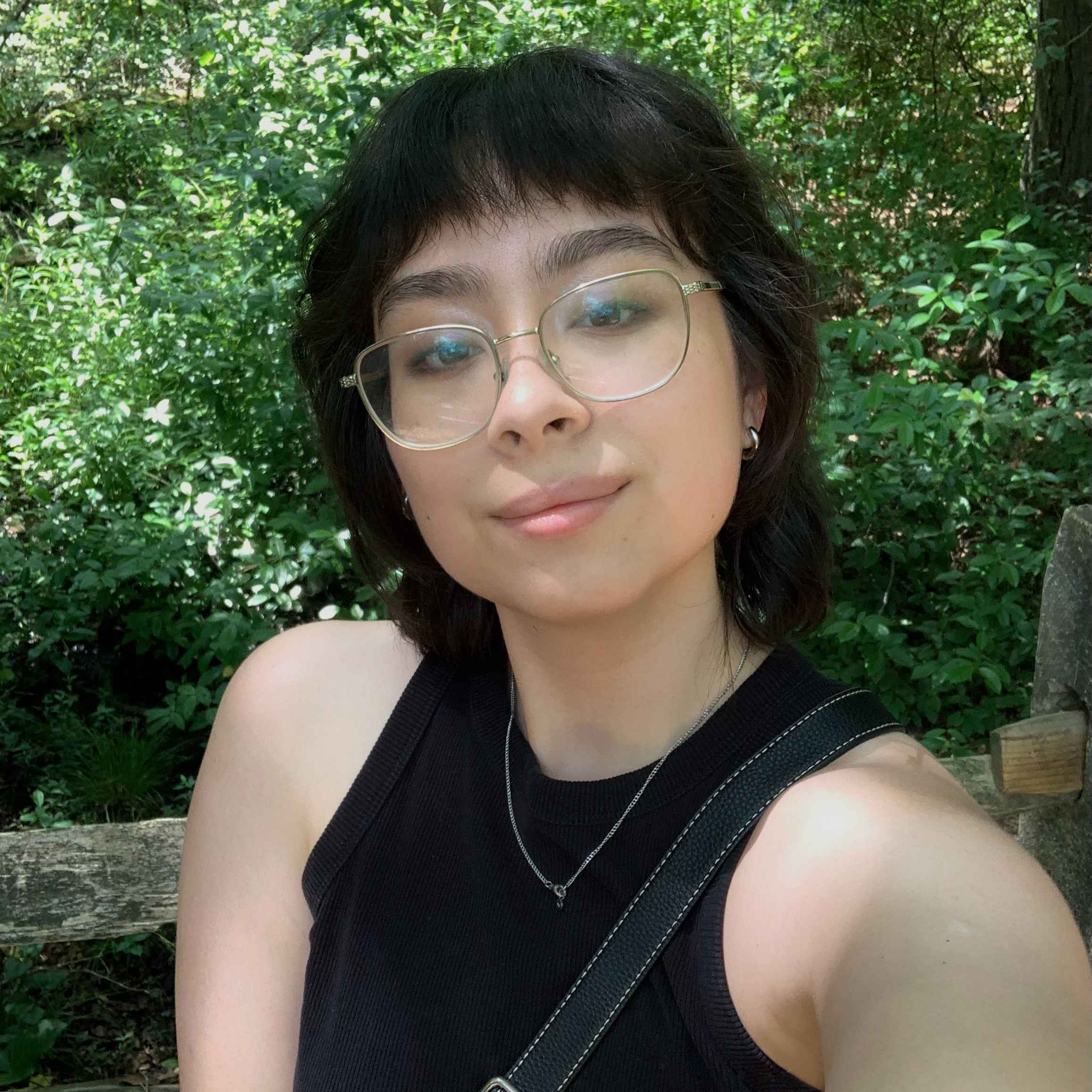 Kira stands outdoors in front of bright green leafy bushes and looks at the camera with a slight smile. She has short black hair, light skin, and wears big round glasses. She is wearing a black tank top.